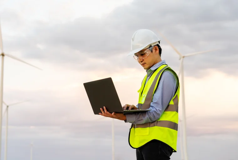 Engineer in white hard hat and safety vest uses a laptop while standing in front of multiple wind turbines