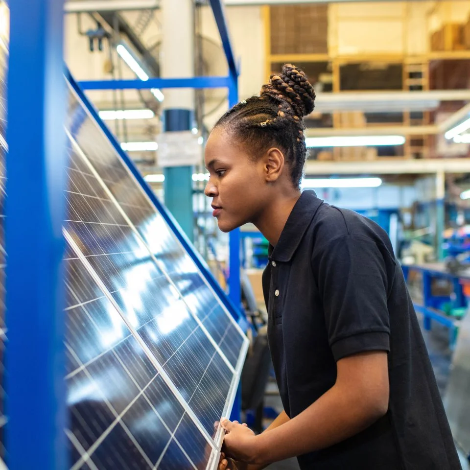 A black woman in a dark polo shirt holds a solar panel in the factory